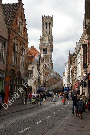 People walk on a narrow street with the tower of The Belfry in the background in the city of Bruges in the province of West Flanders, Belgium.