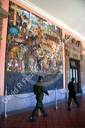 Men in military uniform walk past a mural painted by Diego Rivera at the National Palace in Mexico City, Mexico.