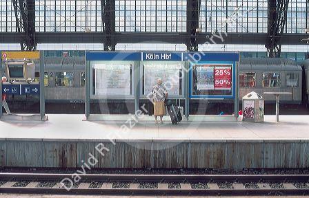 Train station at Cologn, Germany.  In German it is called the Hauptbahnhof.  Elderly woman reads schedule on platform.