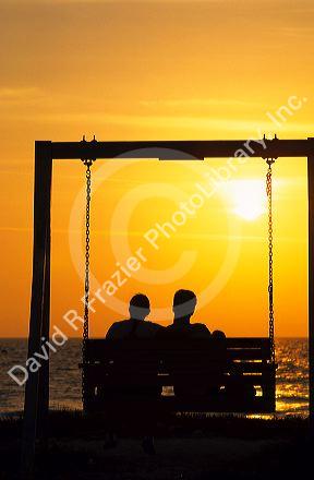 A man and woman sitting on a bench swing silhouetted at sunset.