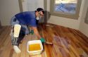 A flooring finisher wearing a respirator applies sealer to a new hard wood floor.