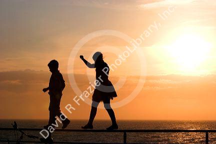 Silhouette of children walking on a railing at sunset by the sea.