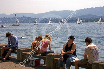 People sit along the Zurichsee Lake with sailboats behind at Zurich, Switzerland.

switzerland, swiss, europe, european, travel, tourism, swiss alps, alps, alpine, zurich, sailboat, sail boat, boat, zurichsee lake, zurichsee, lake