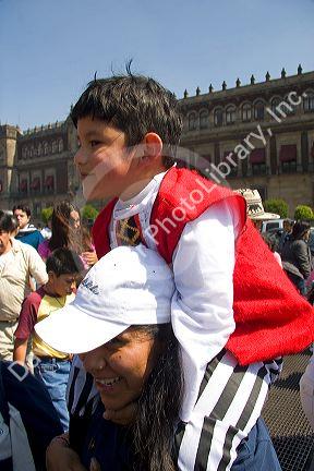 A young Mexican boy sits on his mothers shoulders to see over a crowd of people celebrating the Day of the Dead in the Zocalo in Mexico City, Mexico.