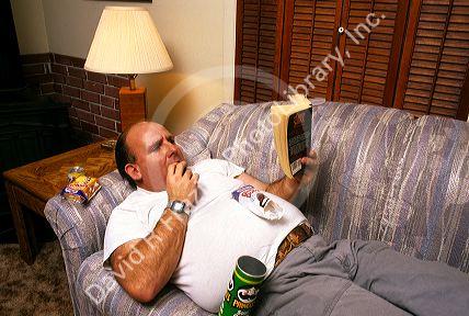 Overweight man laying on a couch eating and reading a book. MR