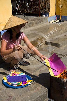 Vietnamese woman burning colorful paper to honor departed ancestors during Tet in Hoi An, Vietnam.
