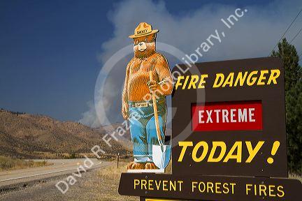 Smokey Bear extreme fire danger sign with a plume of smoke from a forest fire in the back ground in Boise County, Idaho, USA.