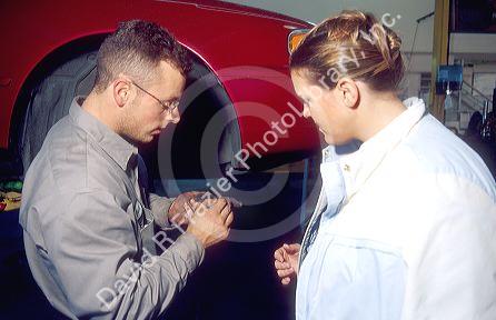 A male adult auto mechanic shows a female adult customer a worn out brake pad from her car.