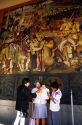 A mexican family stands in front of a Diego Rivera mural at the National Palace in Mexico City, Mexico.