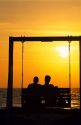 A man and woman sitting on a bench swing silhouetted at sunset.
