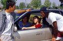 Male teens lean on a car talking to female teens inside. MR