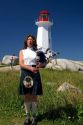 Scottish woman in traditional dress playing the bag pipes in front of a lighthouse at Peggy's Cove, Nova Scotia, Canada.